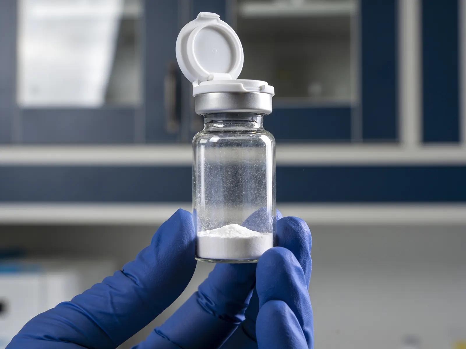 A gloved hand holding a vial of white lyophilised powder